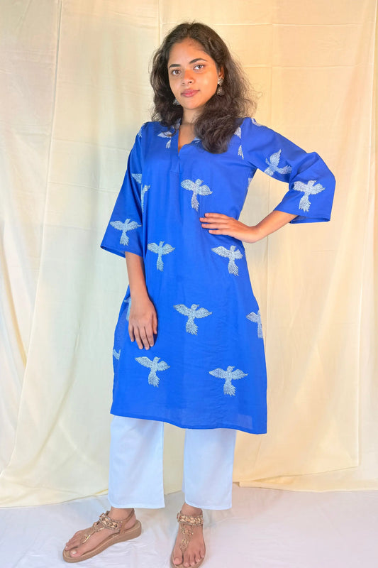Woman wearing a blue kurta with white patterns against a beige curtain background