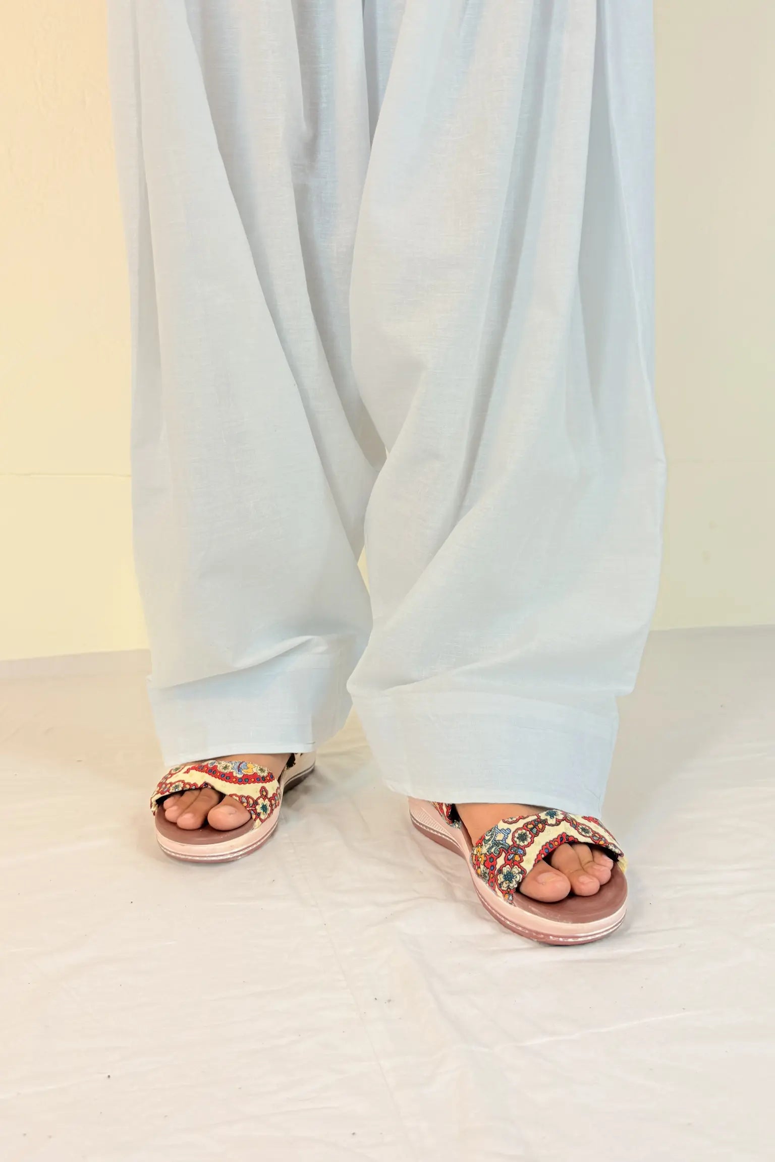 Person wearing light blue pants and pink sandals with decorative straps on a white floor.