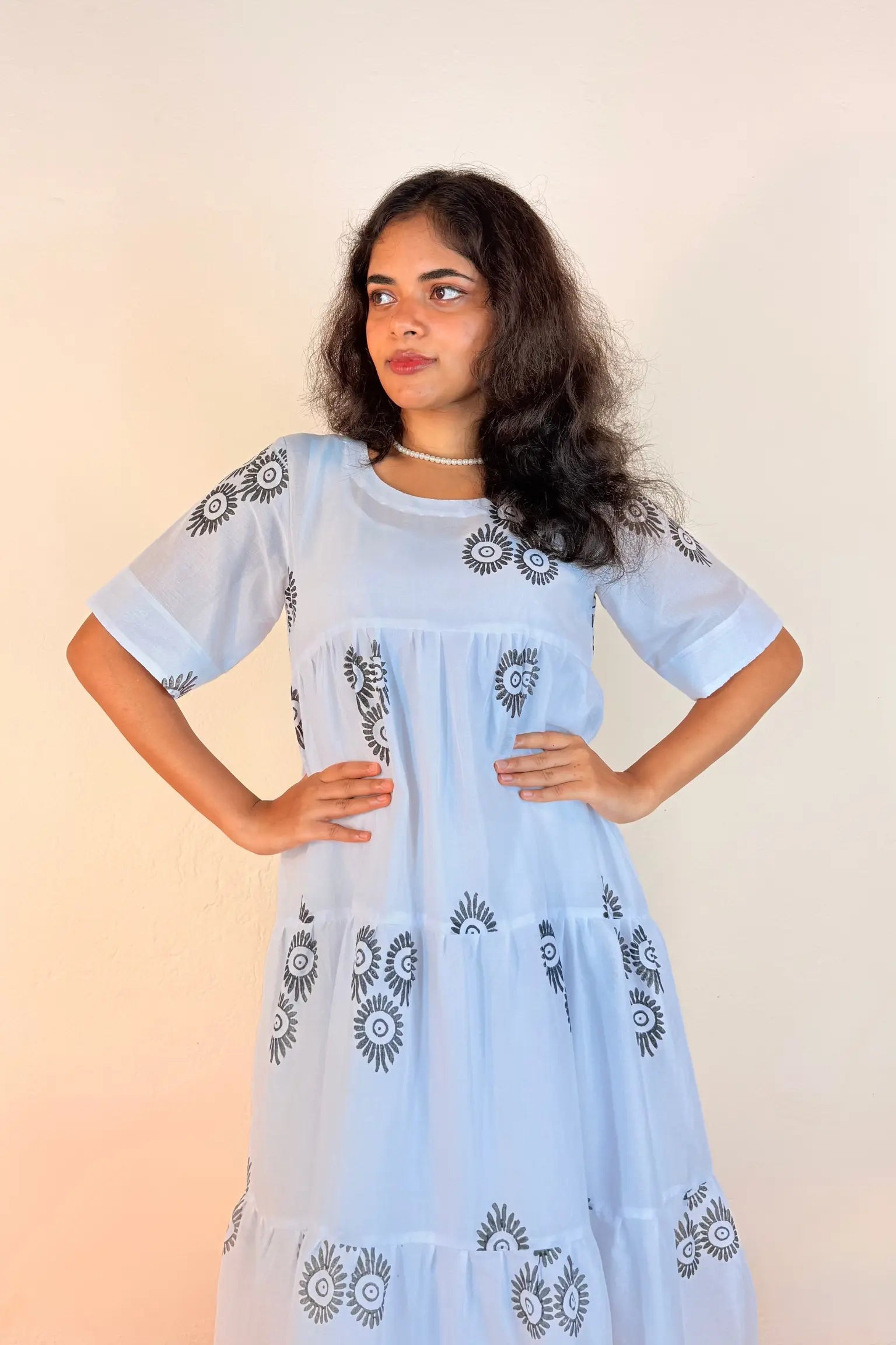 A woman modeling a light blue tiered dress with black floral embroidery, standing against a plain beige background.