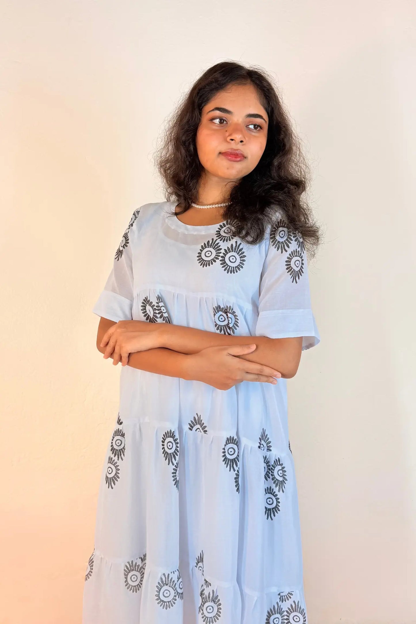 A young woman with dark hair is wearing a light blue tiered dress with black and white sunburst floral patterns, standing against a plain light beige background.