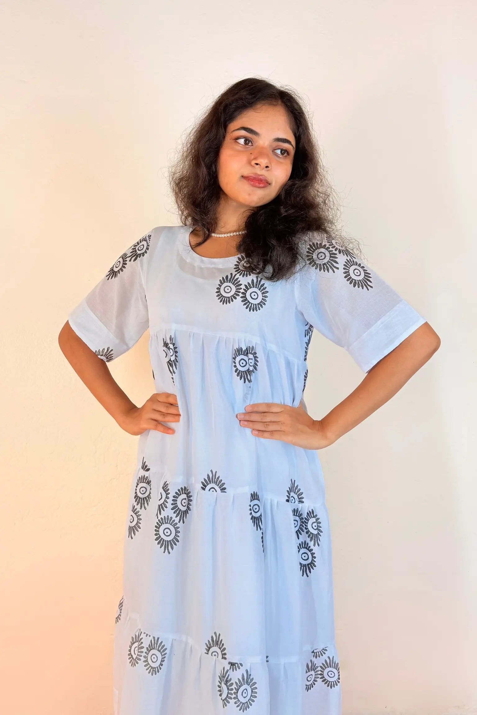 A woman modeling a light blue tiered maxi dress with black and white floral embroidery on the bodice, sleeves, and skirt, set against a plain beige background.