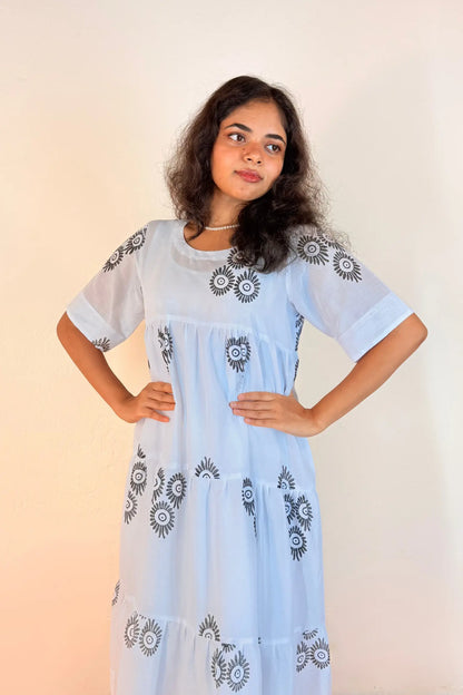 A woman modeling a light blue tiered maxi dress with black and white floral embroidery on the bodice, sleeves, and skirt, set against a plain beige background.