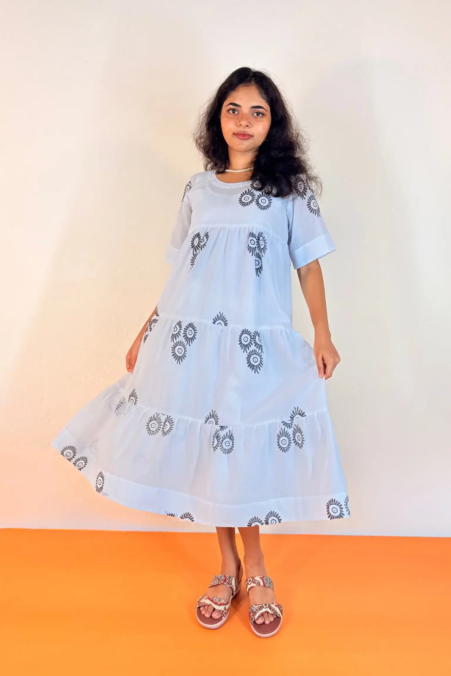 A woman modeling a light blue tiered cotton dress with black and white geometric embroidery, standing against a plain off-white wall with an orange floor.