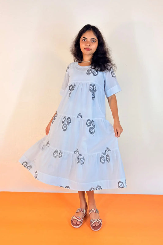 A woman modeling a light blue tiered cotton dress with black and white geometric embroidery, standing against a plain off-white wall with an orange floor.