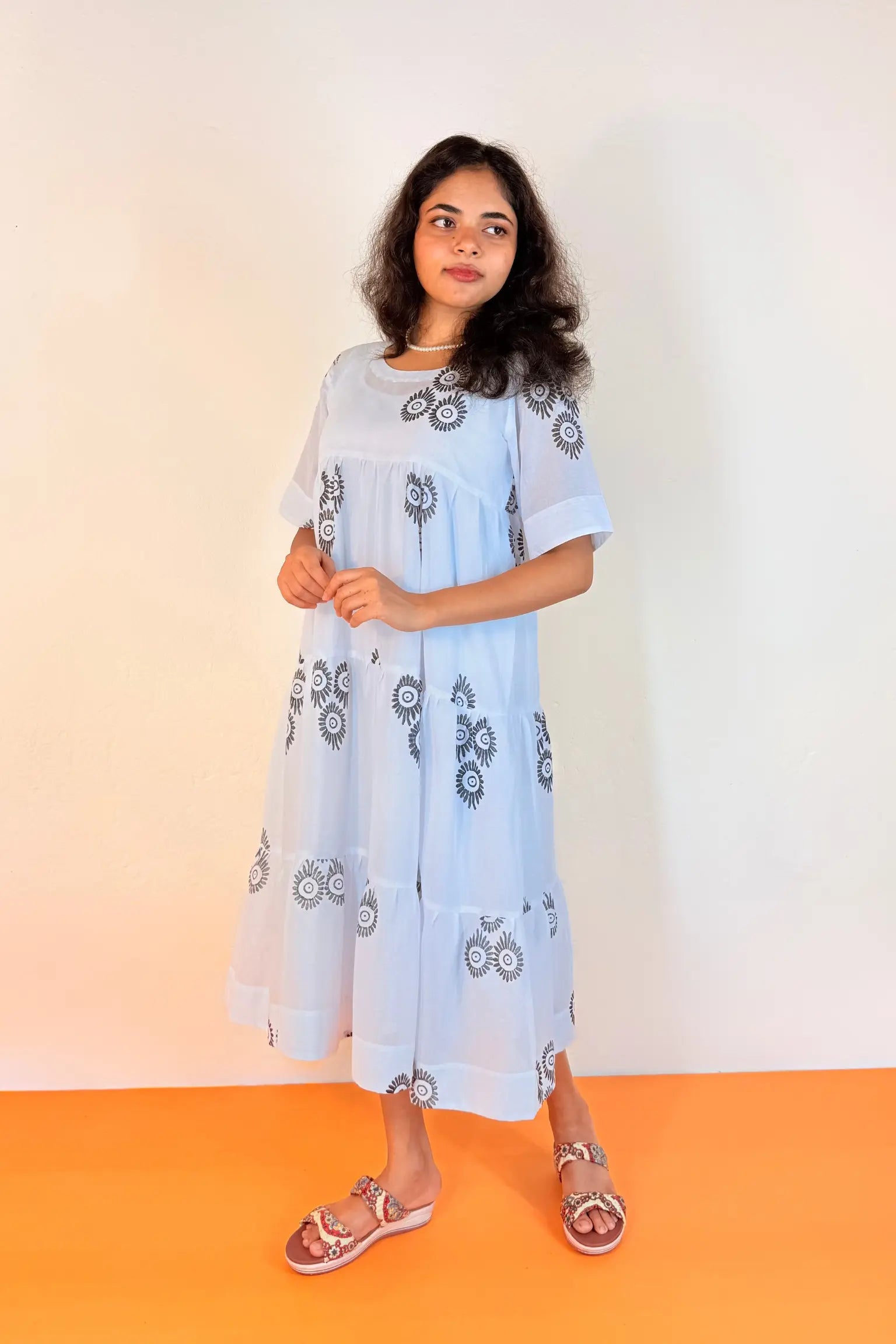 A woman modeling a light blue midi dress with dark gray floral embroidery, standing against a white wall with an orange floor.
