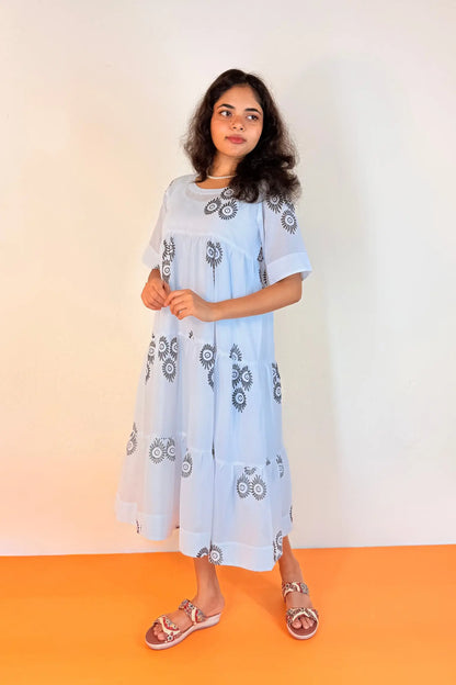 A woman modeling a light blue midi dress with dark gray floral embroidery, standing against a white wall with an orange floor.