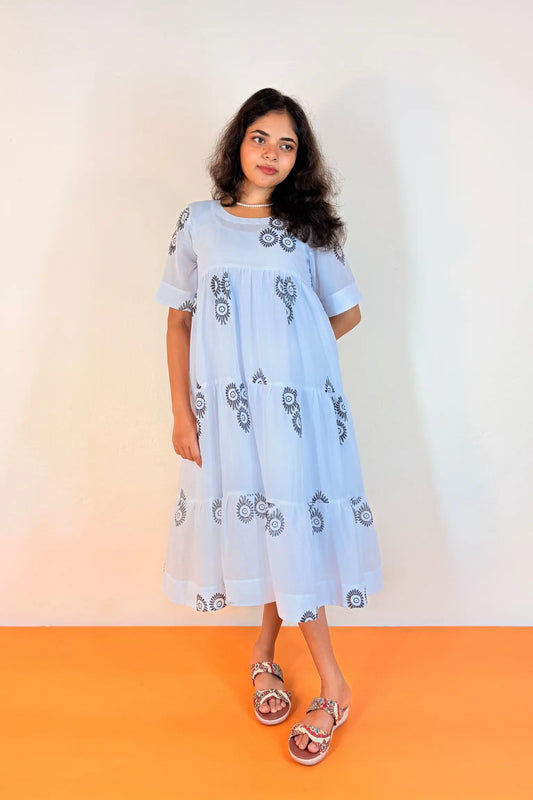 A young girl modeling a light blue short-sleeve midi dress with dark blue circular patterns, standing against a plain off-white wall with an orange floor.