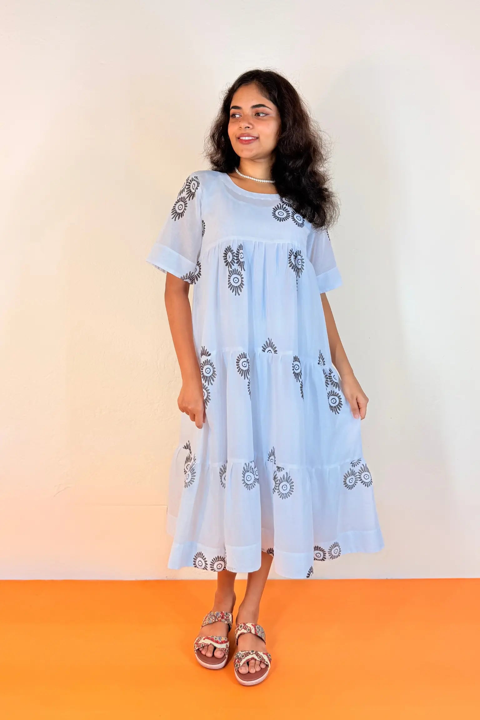 A woman modeling a light blue tiered cotton dress with black printed motifs, standing against a plain off-white wall with an orange floor.