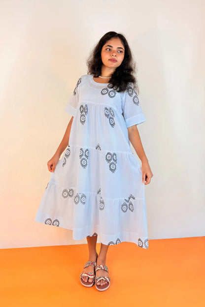 A woman modeling a light blue tiered cotton dress with dark gray block print patterns, paired with brown sandals, standing against a plain beige wall and orange floor.