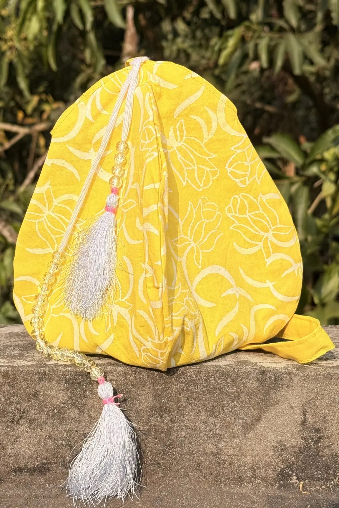 Yellow fabric pouch with floral patterns and tassels on a stone surface with green foliage in the background