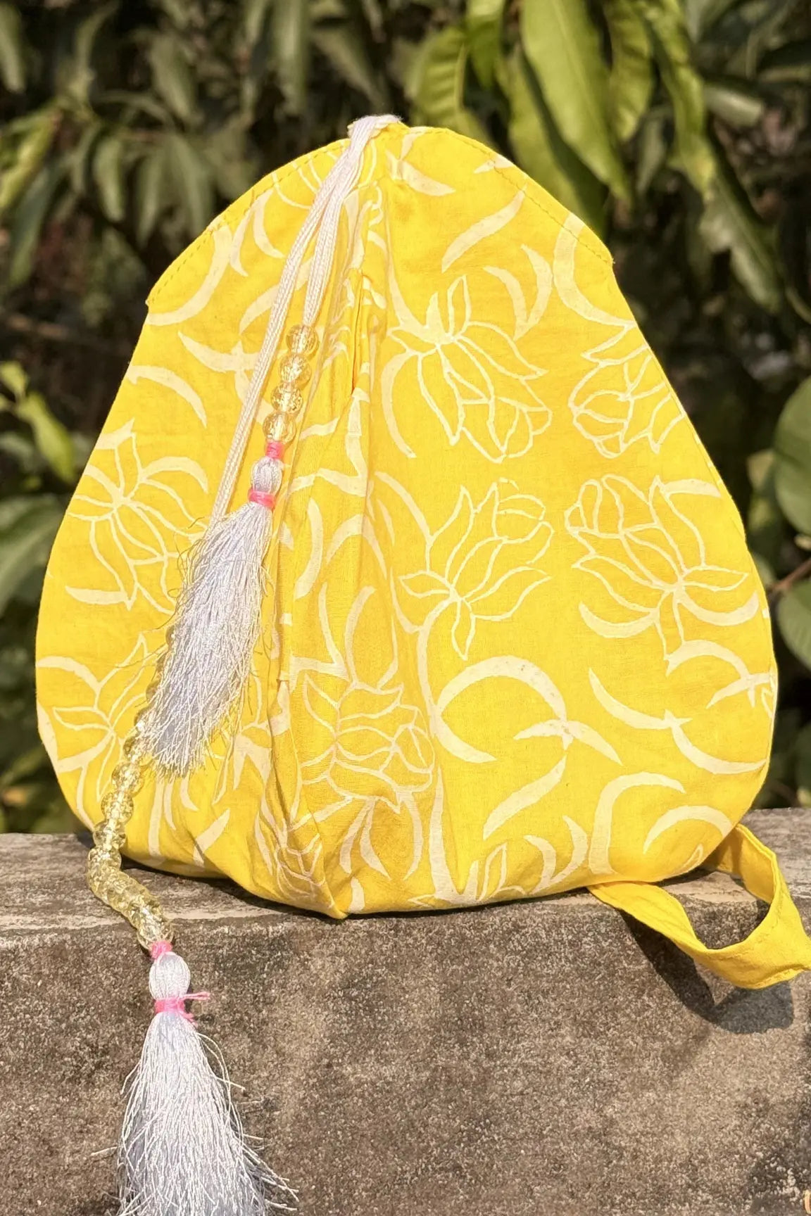 Yellow fabric pouch with floral patterns and tassels on a stone surface with green foliage in the background