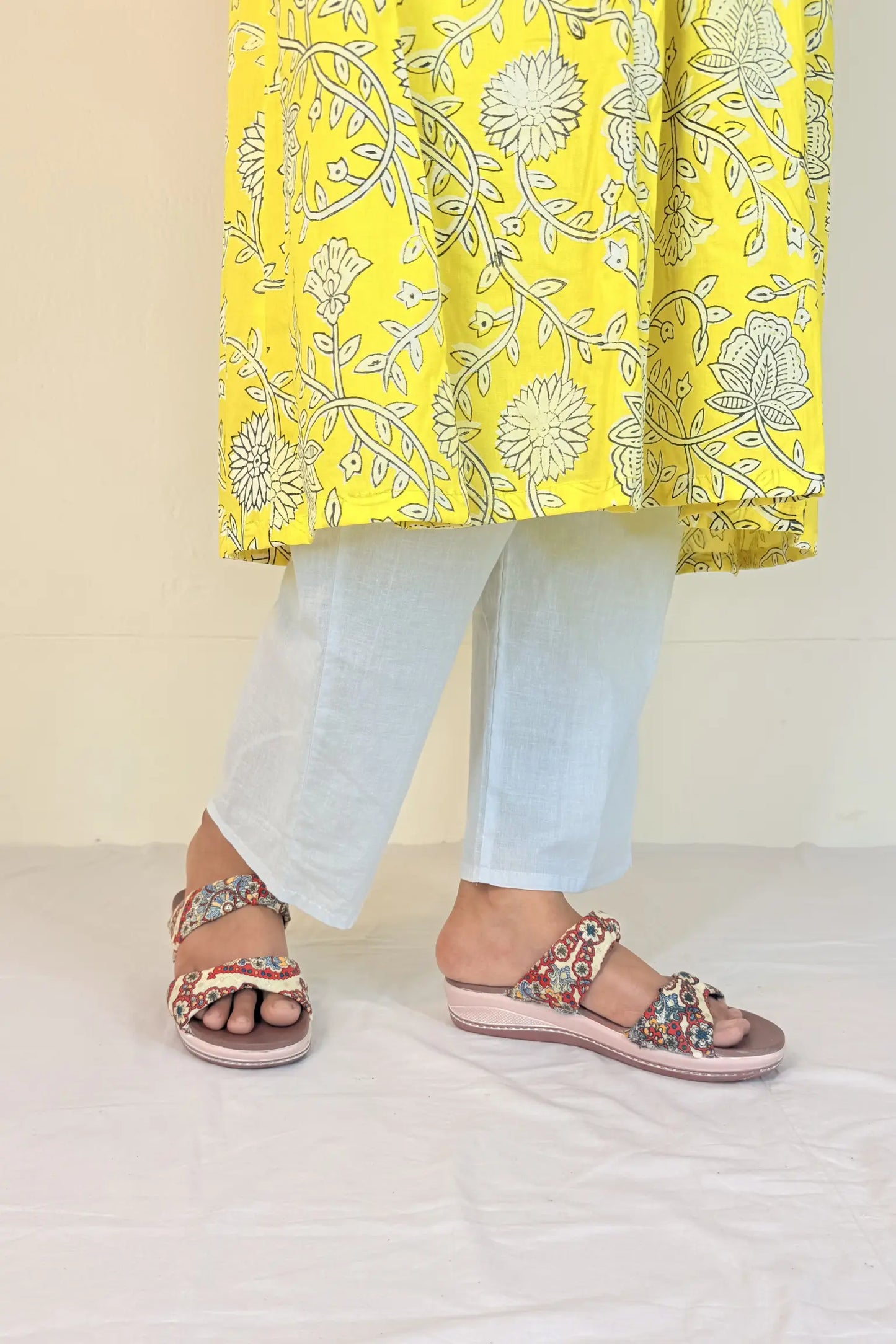 Person wearing a yellow floral kurta with light blue pants and pink sandals on a white floor.