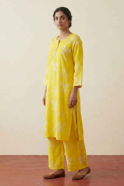 A woman modeling a bright yellow kurta and trouser set with a white floral pattern, standing against a plain beige wall.