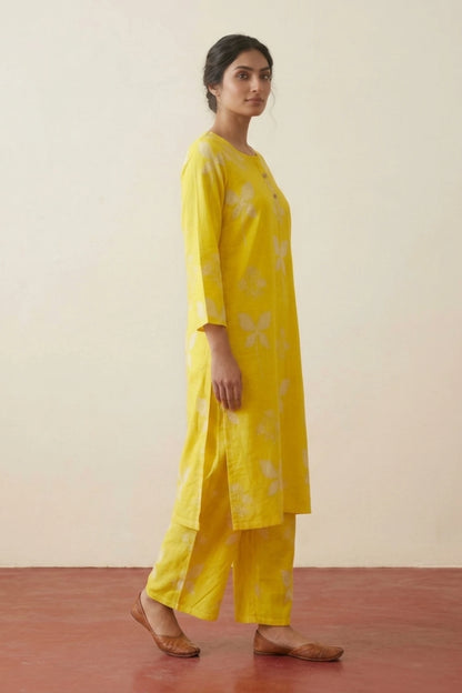 A woman modeling a bright yellow kurta and matching palazzo pants set with white floral prints, standing in profile against a plain beige wall and reddish-brown floor.