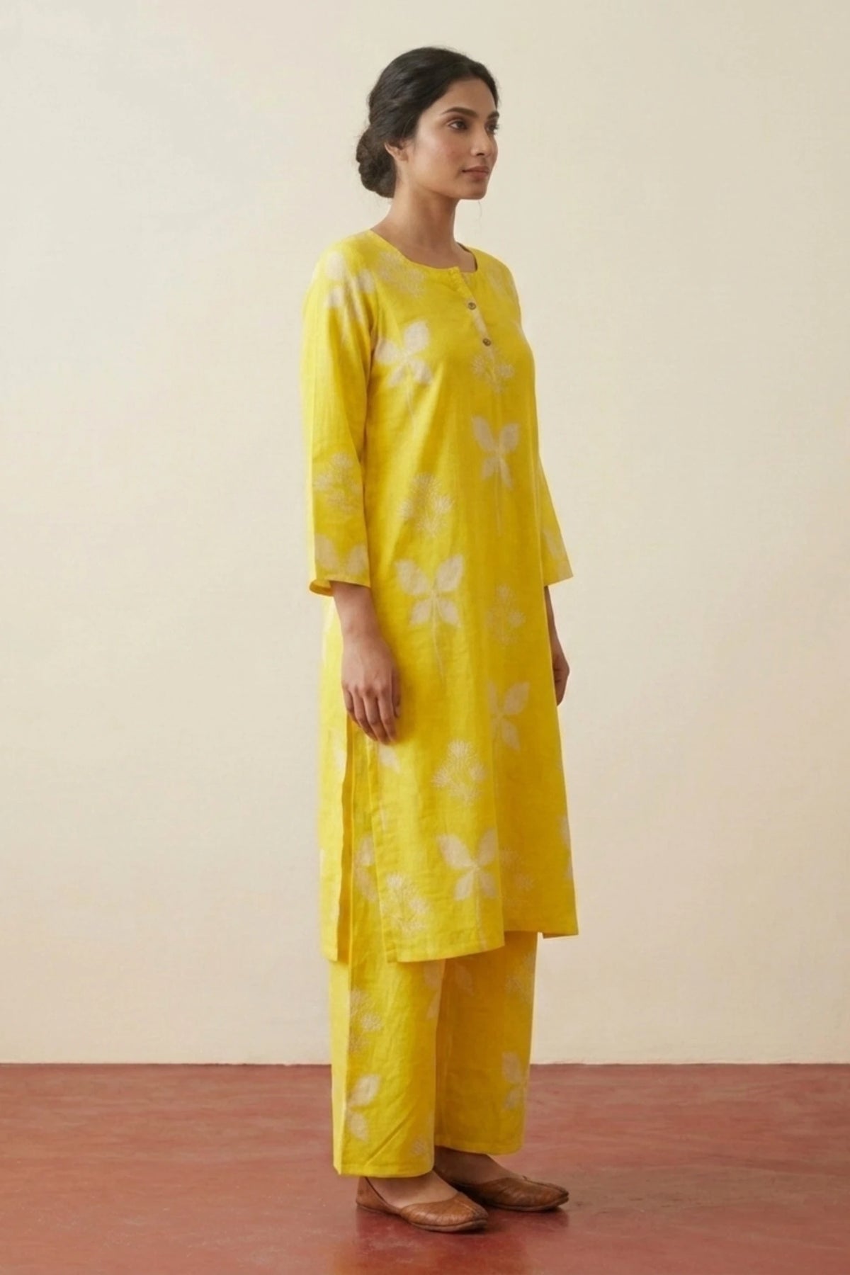 A woman modeling a bright yellow kurta set with white floral motifs, consisting of a long-sleeved tunic and matching wide-leg trousers, set against a plain beige wall and reddish-brown floor.