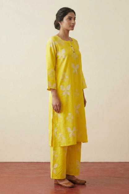 A woman modeling a bright yellow kurta set with white floral motifs, consisting of a long-sleeved tunic and matching wide-leg trousers, set against a plain beige wall and reddish-brown floor.
