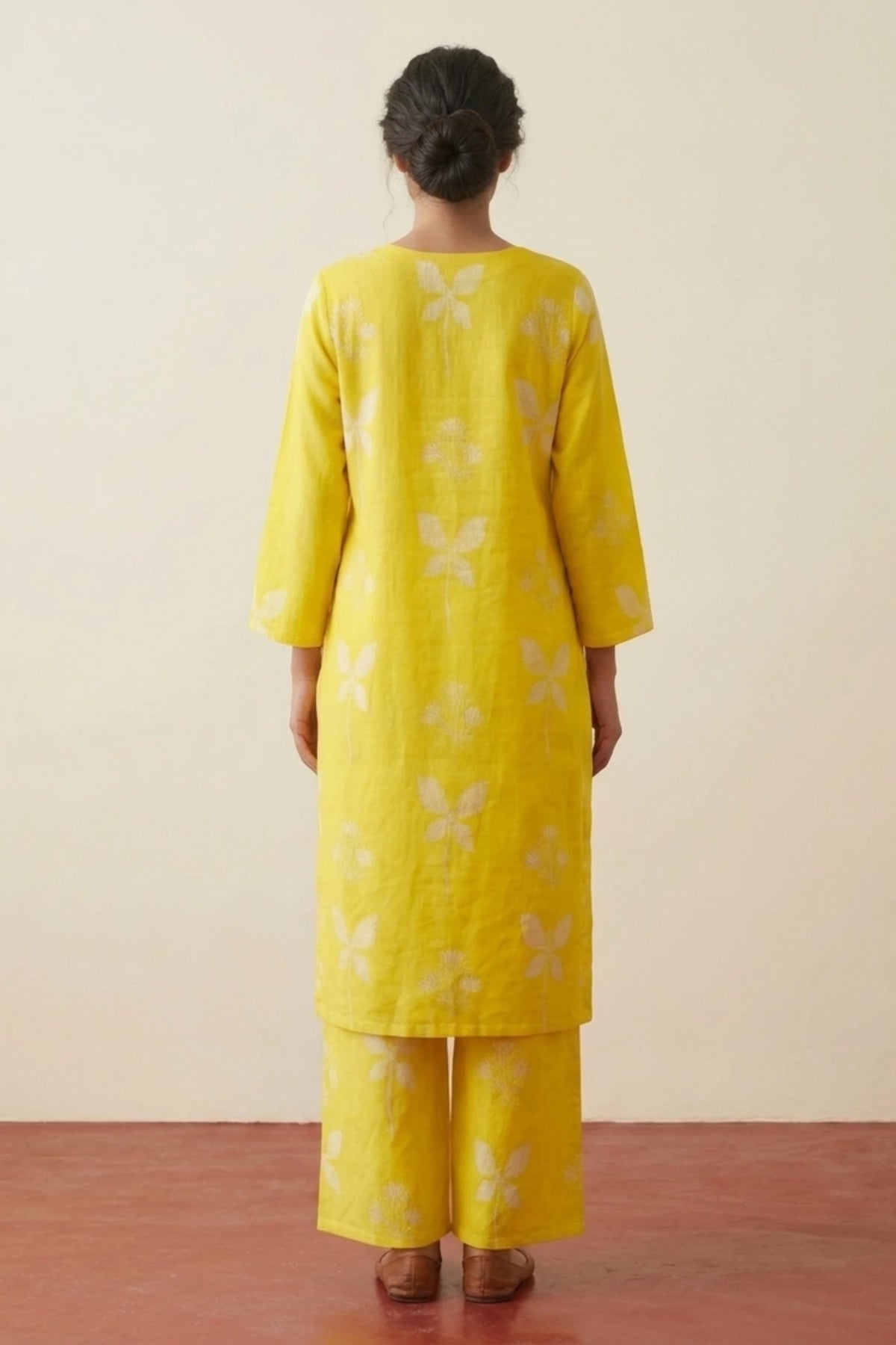 A woman viewed from behind wearing a bright yellow kurta and matching trousers with a white floral block print, standing against a plain beige wall and reddish-brown floor.