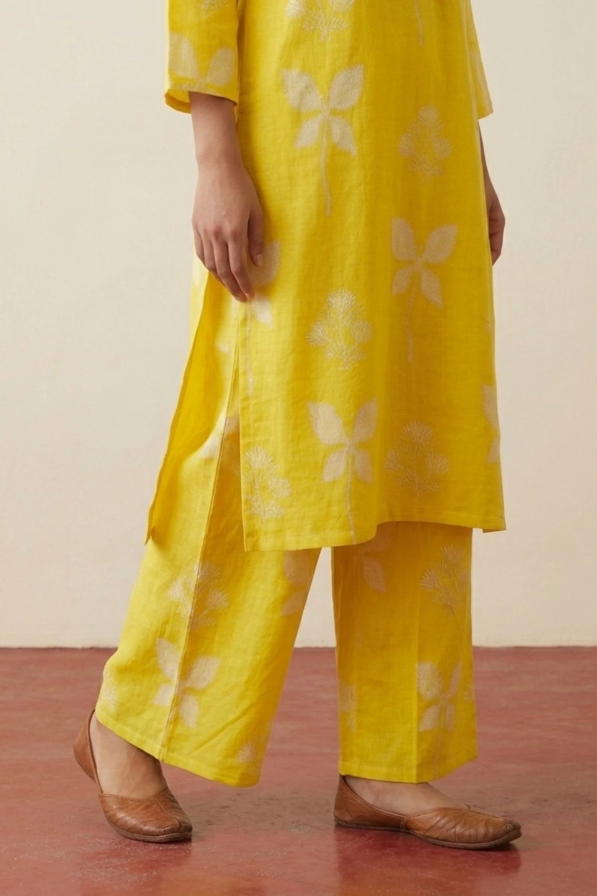A person wearing a bright yellow kurta and matching wide-leg pants with a white floral print, paired with brown leather shoes, standing against a plain beige wall and reddish-brown floor.