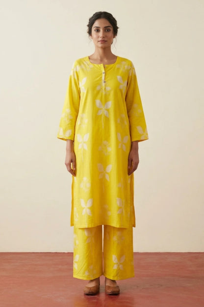 A woman modeling a bright yellow three-piece kurta set with white floral motifs, standing against a plain beige wall and reddish-brown floor.