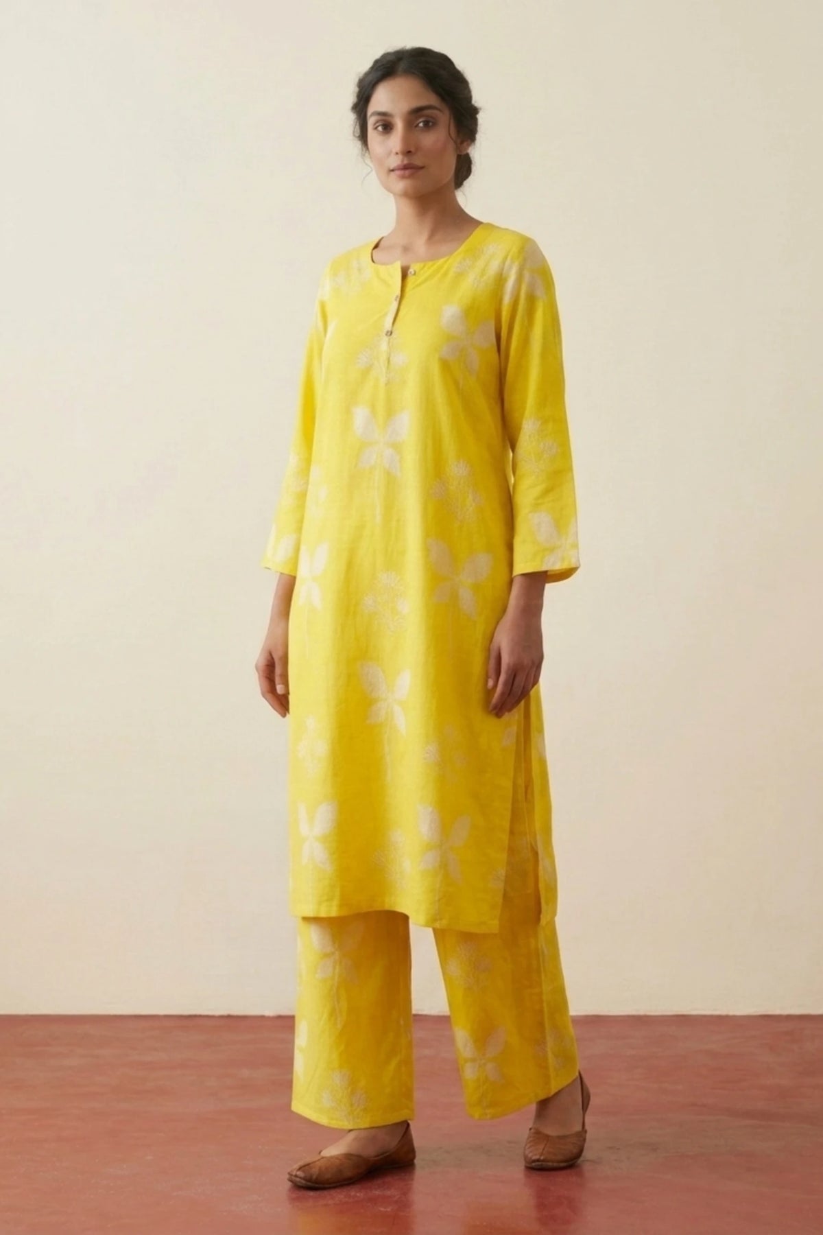 A woman modeling a bright yellow kurta set with white floral print, consisting of a long-sleeve tunic and matching wide-leg pants, set against a plain beige wall and reddish-brown floor.