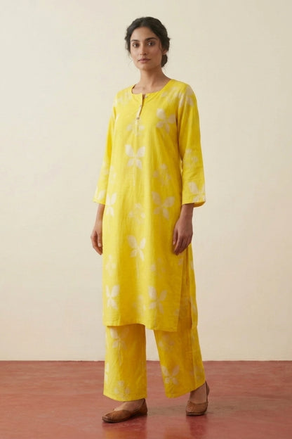 A woman modeling a bright yellow kurta set with white floral print, consisting of a long-sleeve tunic and matching wide-leg pants, set against a plain beige wall and reddish-brown floor.