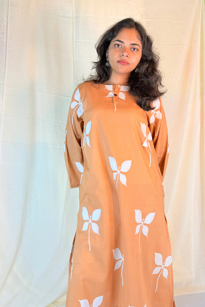 Woman wearing a long, light brown dress with white floral patterns against a plain background