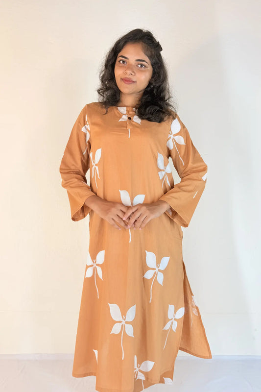 Woman wearing a long, light brown dress with white floral patterns against a plain background