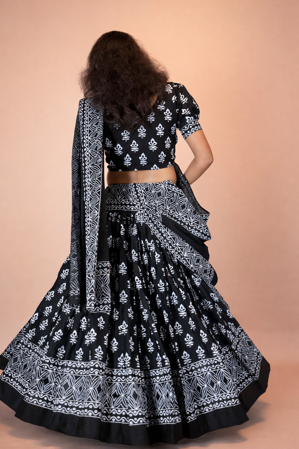 A woman seen from the back wearing a black and white handcrafted block print cotton gopi dress with intricate traditional motifs, flared skirt, and matching dupatta, photographed against a soft warm peach studio background.