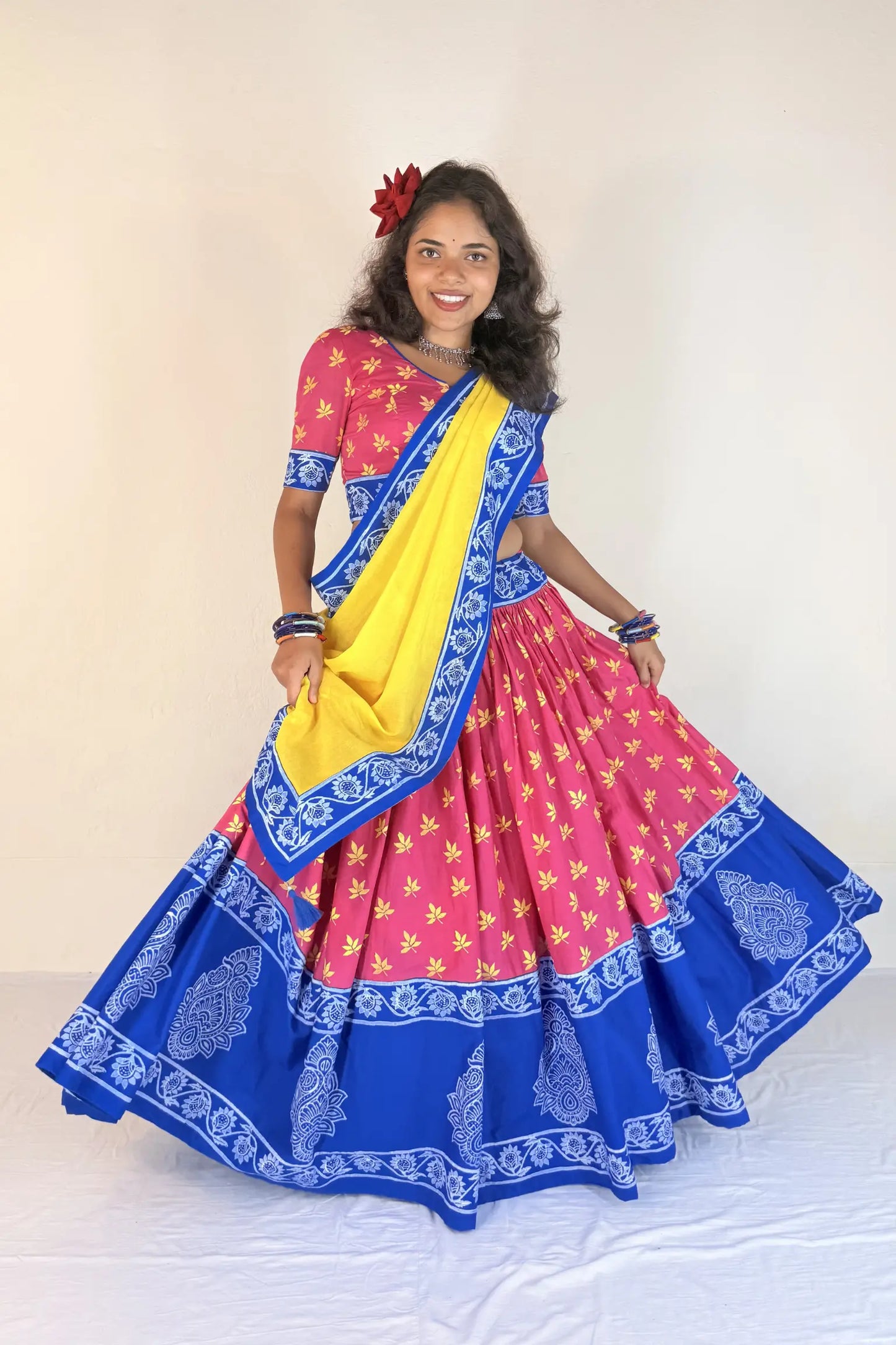 Woman wearing a colorful traditional outfit with a pink blouse, blue and yellow saree, and floral hair accessory.