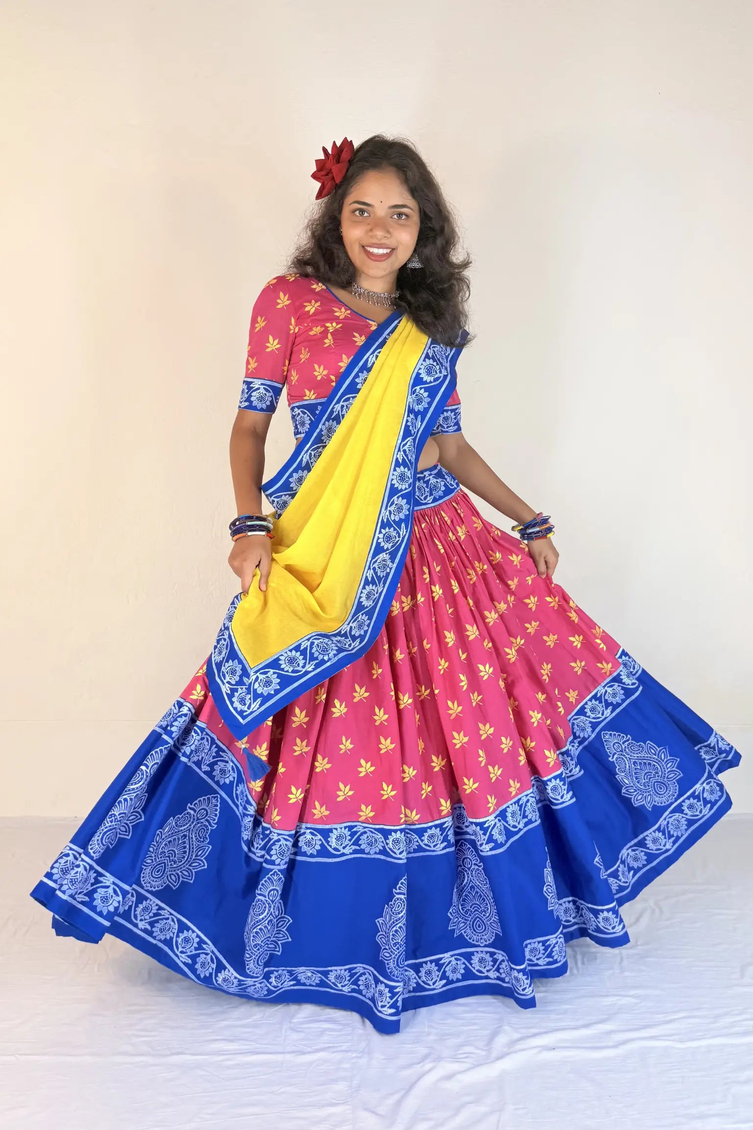 Woman wearing a colorful traditional outfit with a pink blouse, blue and yellow saree, and floral hair accessory.