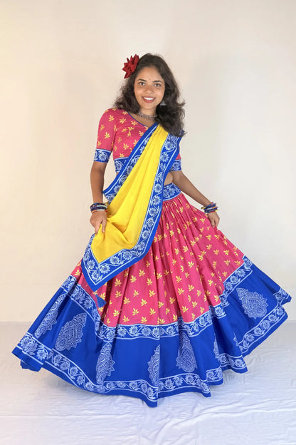 Woman wearing a colorful traditional outfit with a pink blouse, blue and yellow saree, and floral hair accessory.