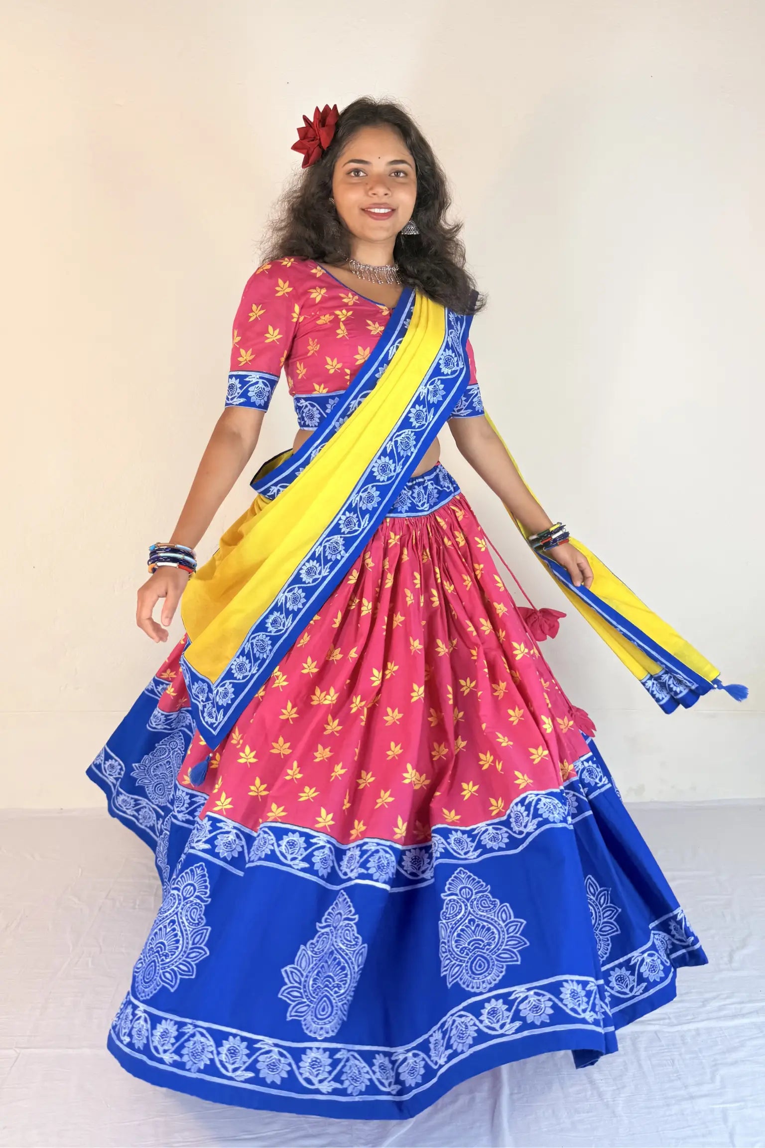 Woman wearing a colorful traditional outfit with a pink blouse, blue saree, and yellow sash against a plain background.