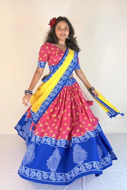 Woman wearing a colorful traditional outfit with a pink blouse, blue saree, and yellow sash against a plain background.