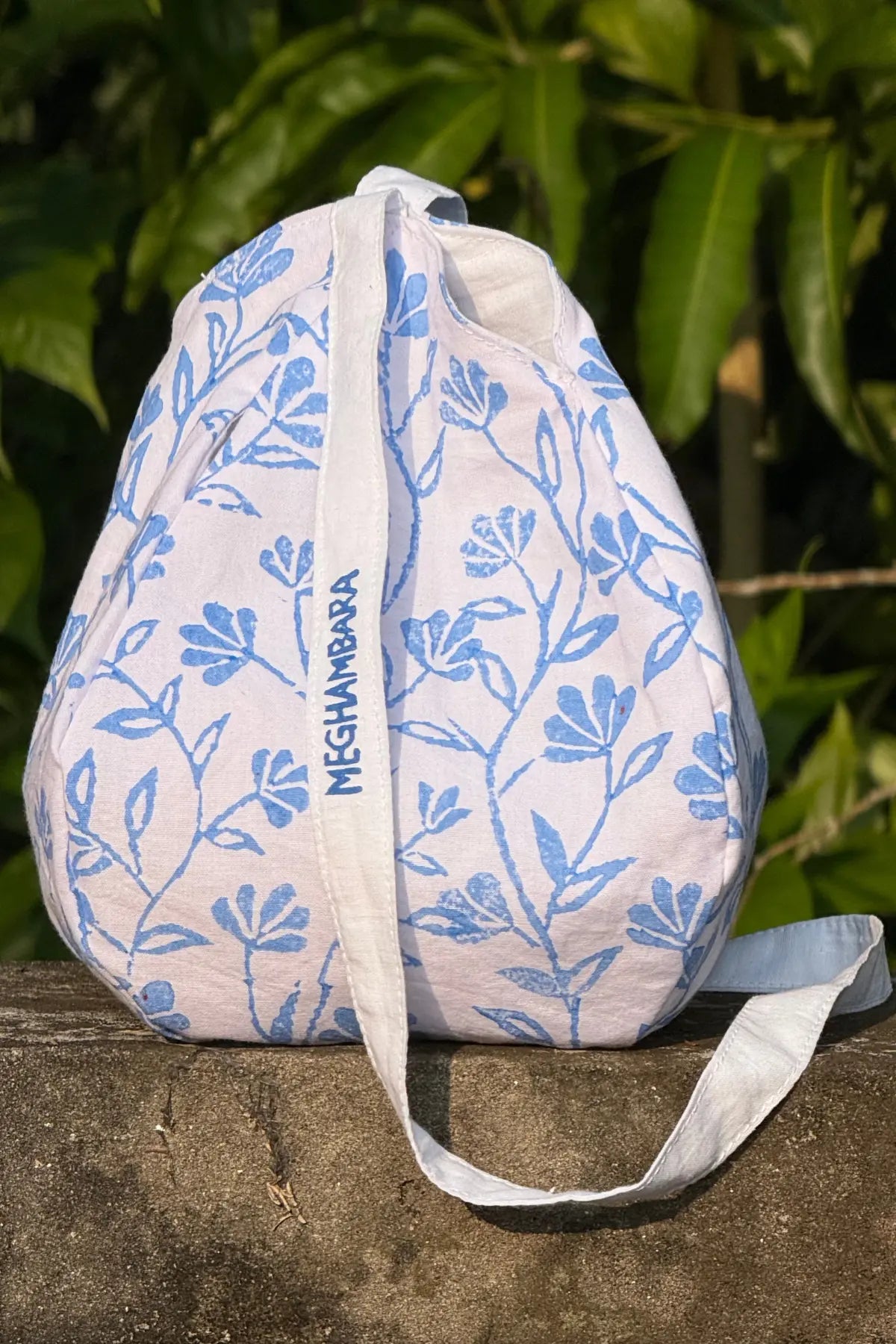 White cotton japa mala bag with blue hand block floral print, front view showing Meghambara branding strap, photographed outdoors
